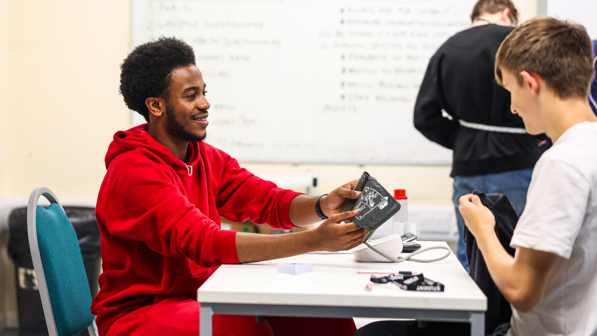 Student in a classroom setting demonstrating a blood pressure monitor to another student across a table, with a whiteboard and additional learning materials visible in the background.