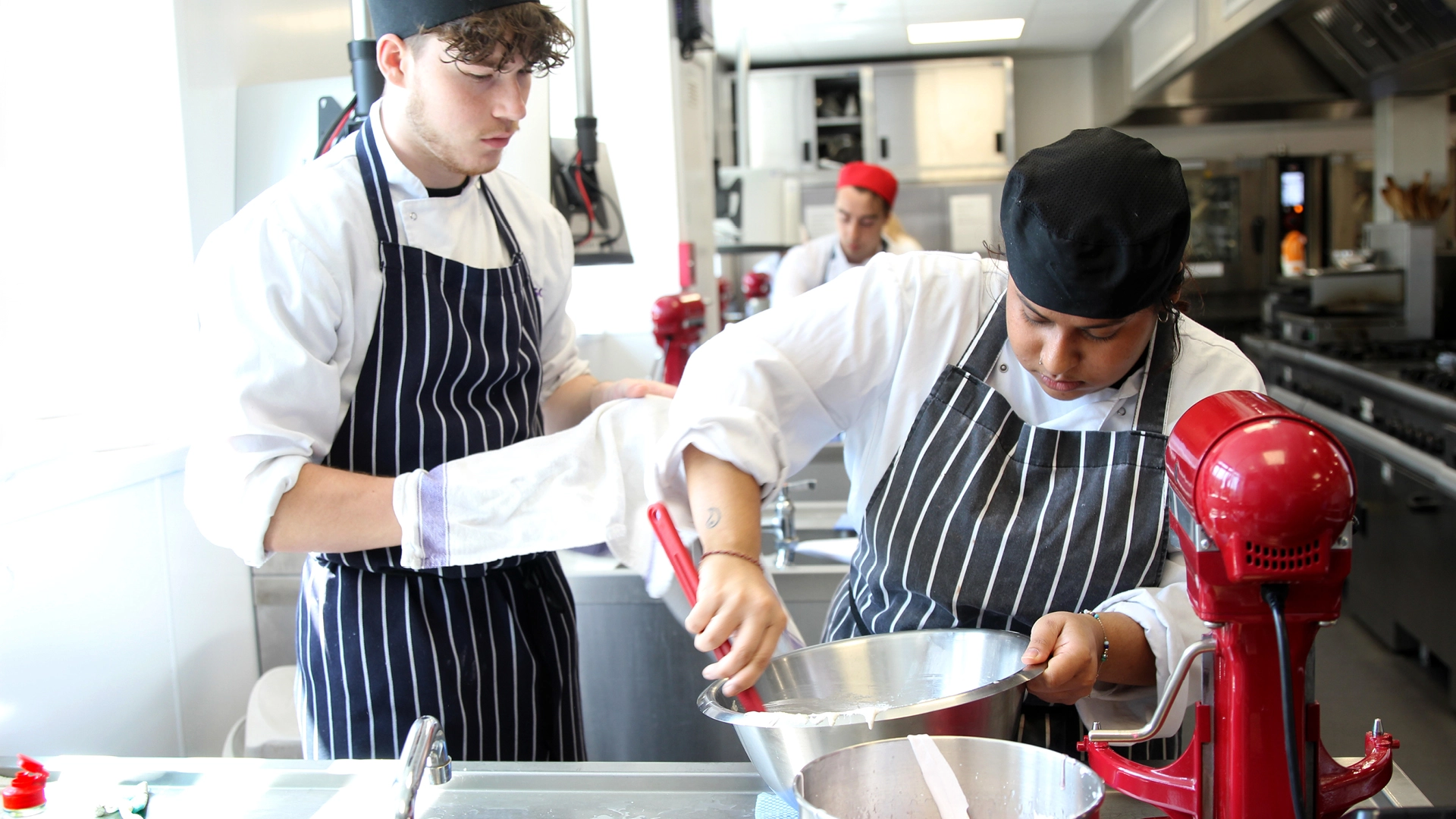 Two Nescot students in chef uniforms work together in a training kitchen, stirring ingredients in a metal bowl next to a red stand mixer, with another student in the background.
