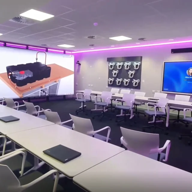 A contemporary teaching space with rows of desks and wheeled chairs, featuring large wall-mounted screens and ambient LED lighting for presentations and collaborative learning.