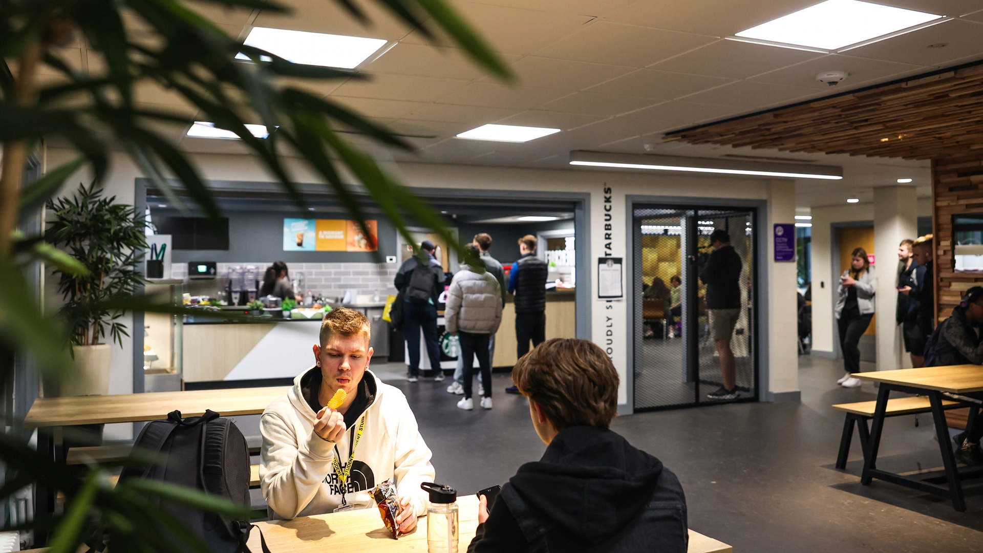 Nescot students sit and chat at tables in a college café while others queue at a food counter in the background.