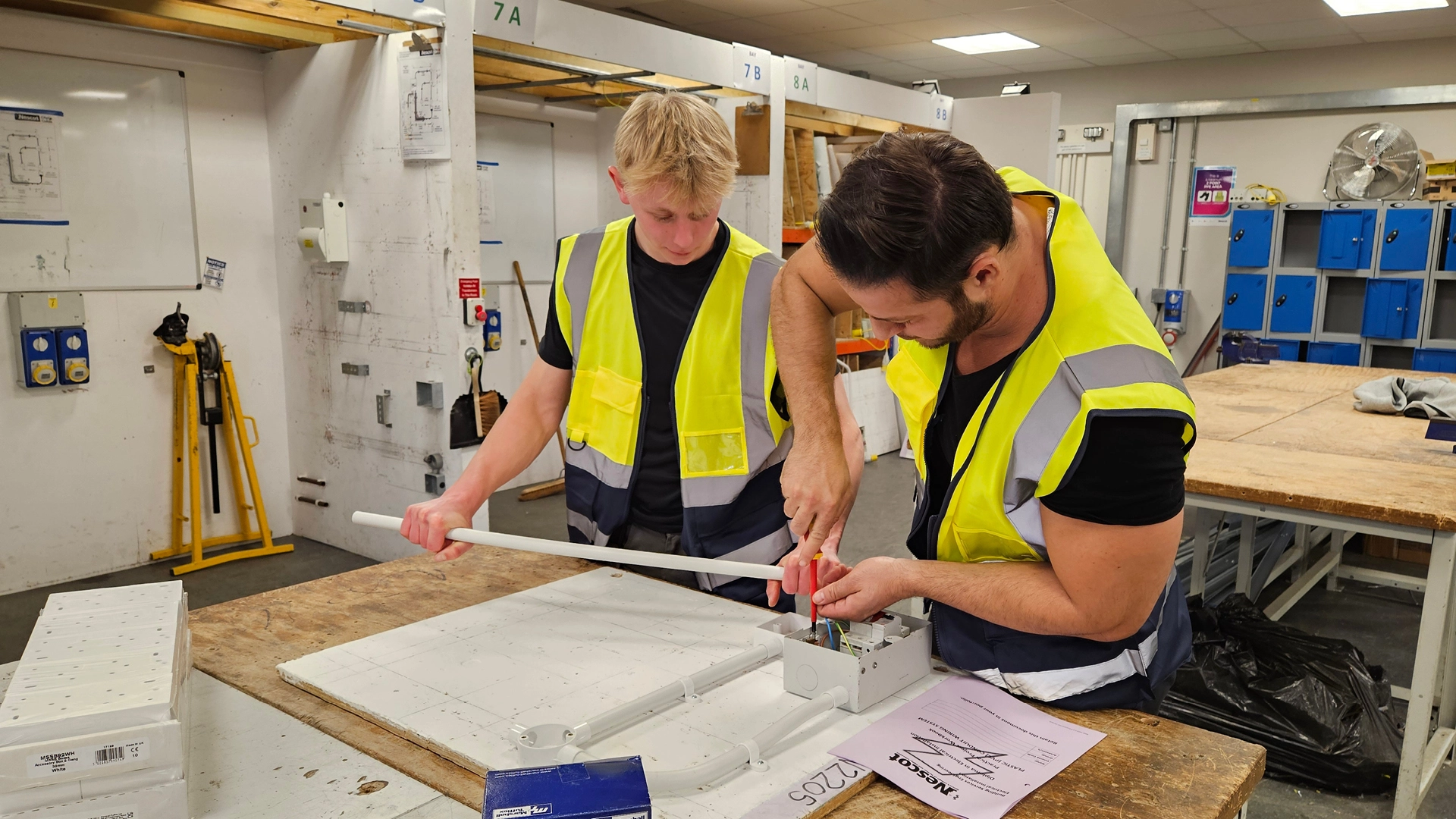 Two Nescot students wearing high-visibility vests and safety gear work together on an electrical wiring task at a workshop bench, using tools and conduit.
