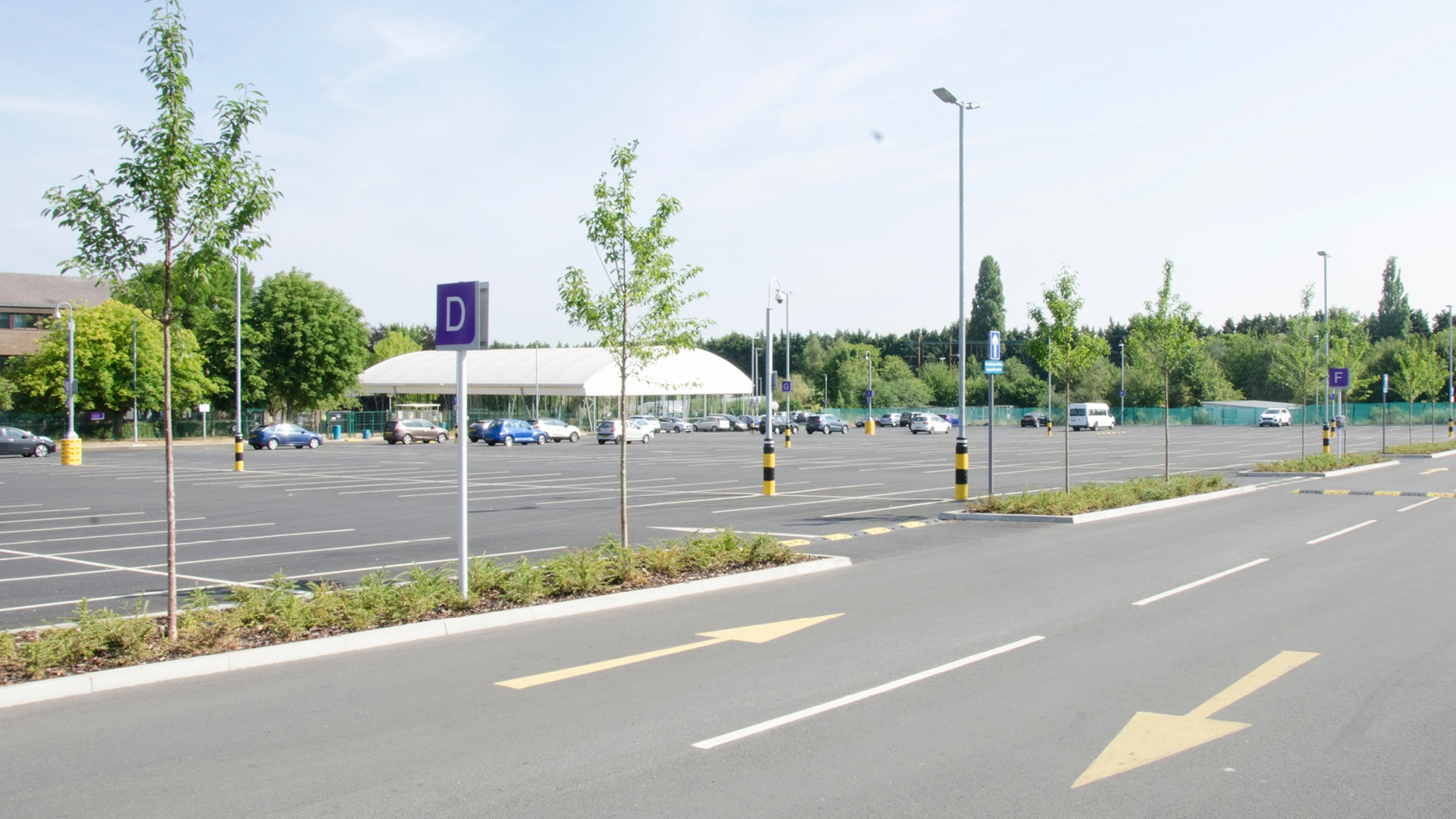 A spacious campus car park with marked bays, directional arrows, and signposts, surrounded by trees and low buildings in the distance.