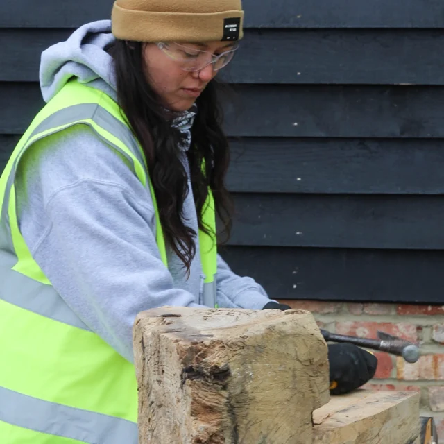 Student wearing high-visibility vest and safety glasses chiselling wood outdoors near a brick wall.