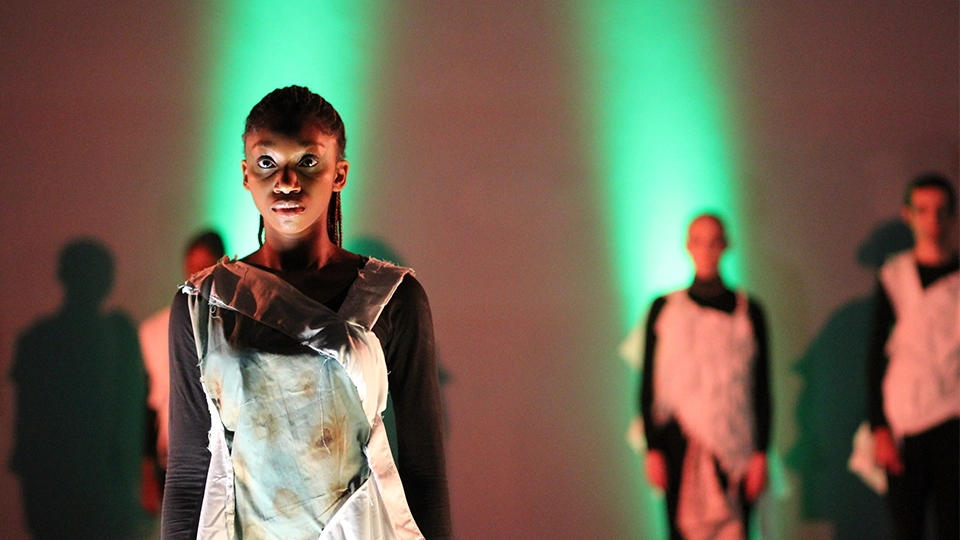 Students modelling fashion pieces under coloured lighting, with others standing behind in a studio setting.