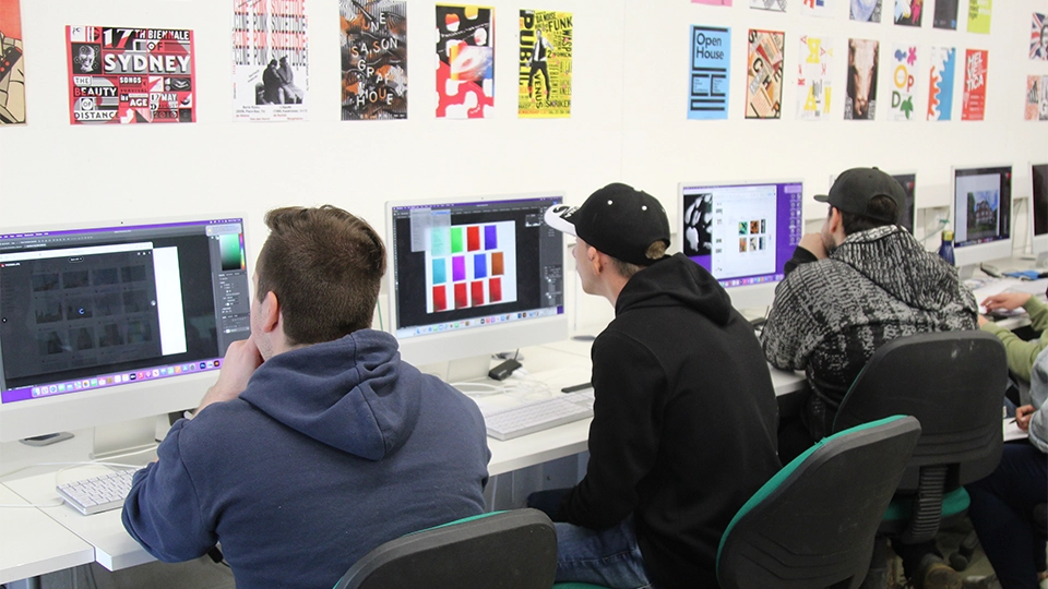 Students seated at desktop computers working on graphic design projects in a classroom.