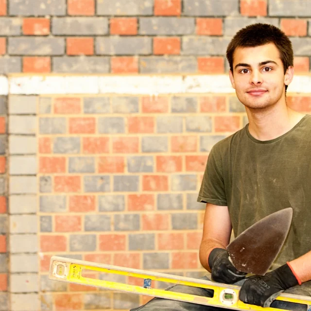 Person holding a trowel and spirit level in front of a brick wall.