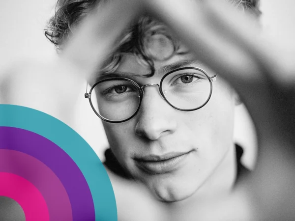 Black and white close-up portrait of a person wearing round glasses, framed by blurred foreground shapes, with a colourful graphic overlay in the lower corner.