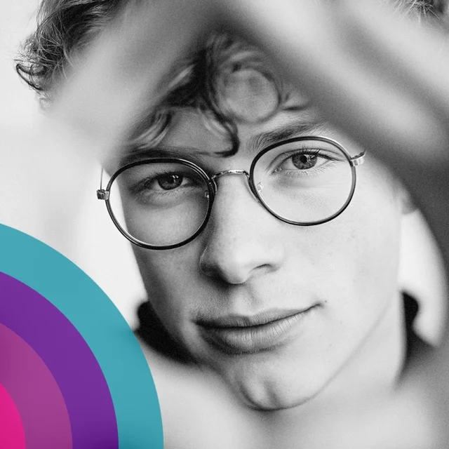 Black and white close-up portrait of a person wearing round glasses, framed by blurred foreground shapes, with a colourful graphic overlay in the lower corner.