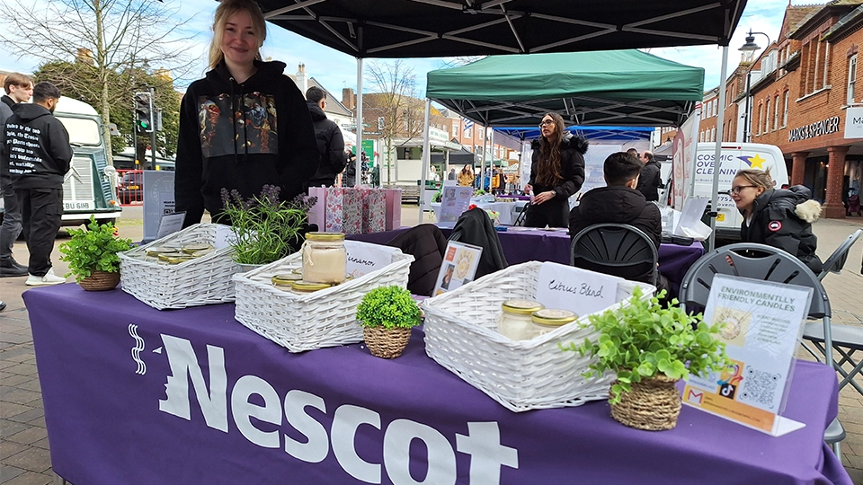 Student standing behind a Nescot-branded stall selling items at an outdoor market.