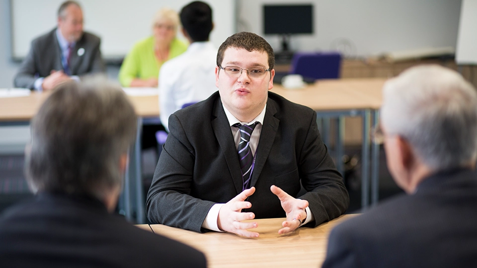 Student in a suit sitting at a table speaking with two people in a classroom setting.