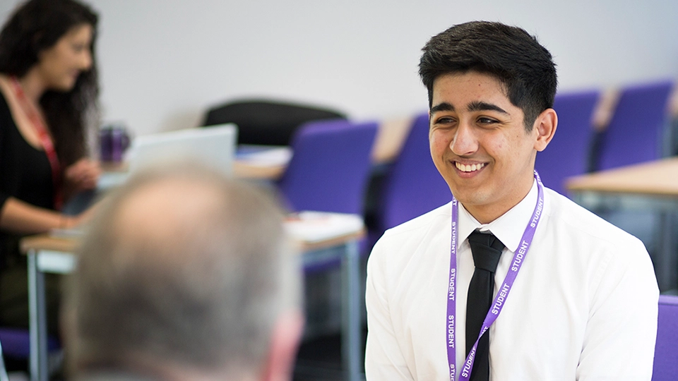 Student wearing a shirt and lanyard speaking with someone across a table in a classroom.