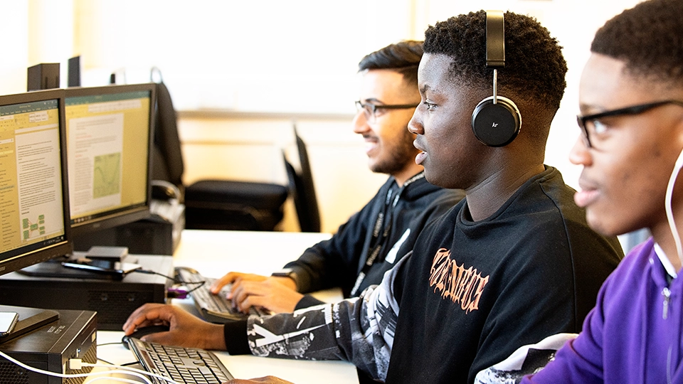 Three students seated at computers working on screens in an IT classroom.