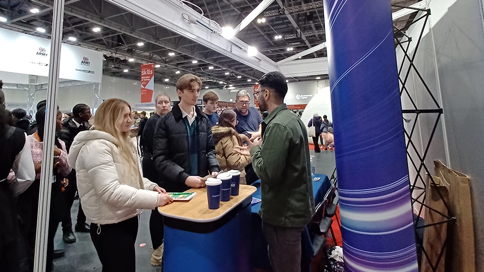 Students speaking with a representative at a stand in a busy exhibition hall.