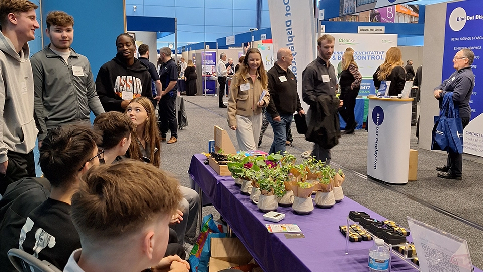 Students gathered around a table displaying potted plants at an indoor event.