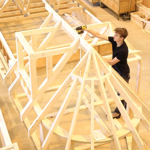 Person using a power drill to assemble a large wooden roof‑frame structure inside a construction workshop, with timber frameworks and tools visible around the space