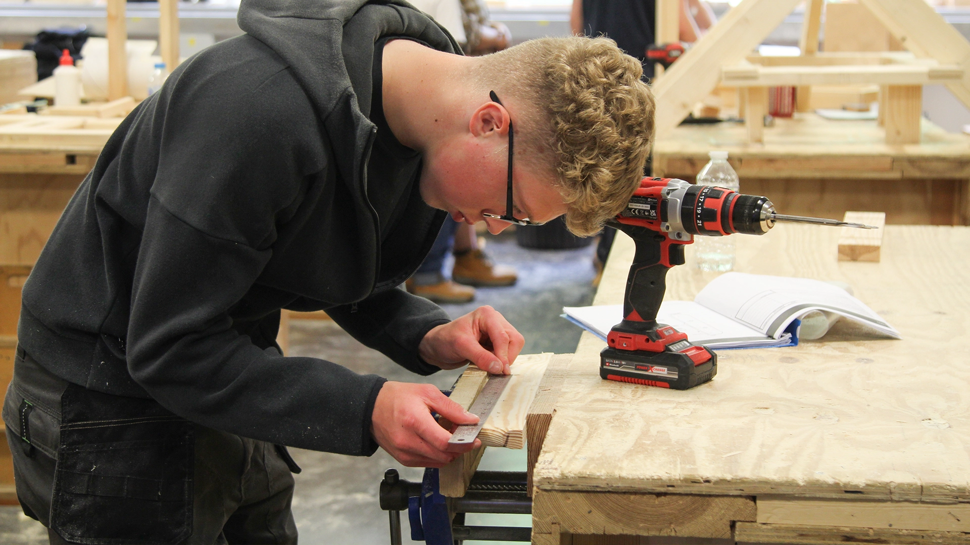 Person measuring a piece of wood on a workbench using a metal ruler, with drill and materials nearby.