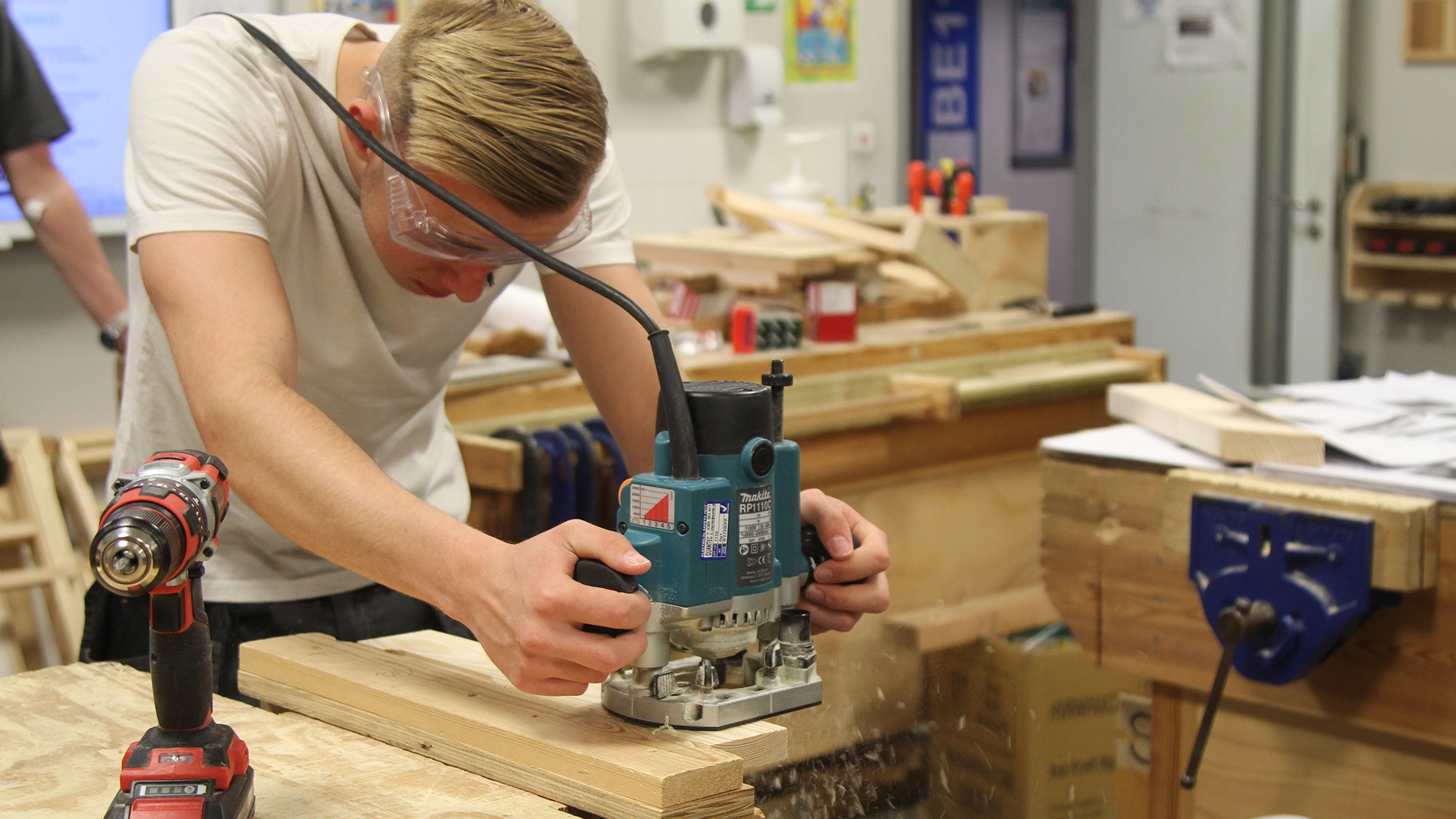 Person using a handheld router to shape a wooden board in a workshop, with a drill on the bench.