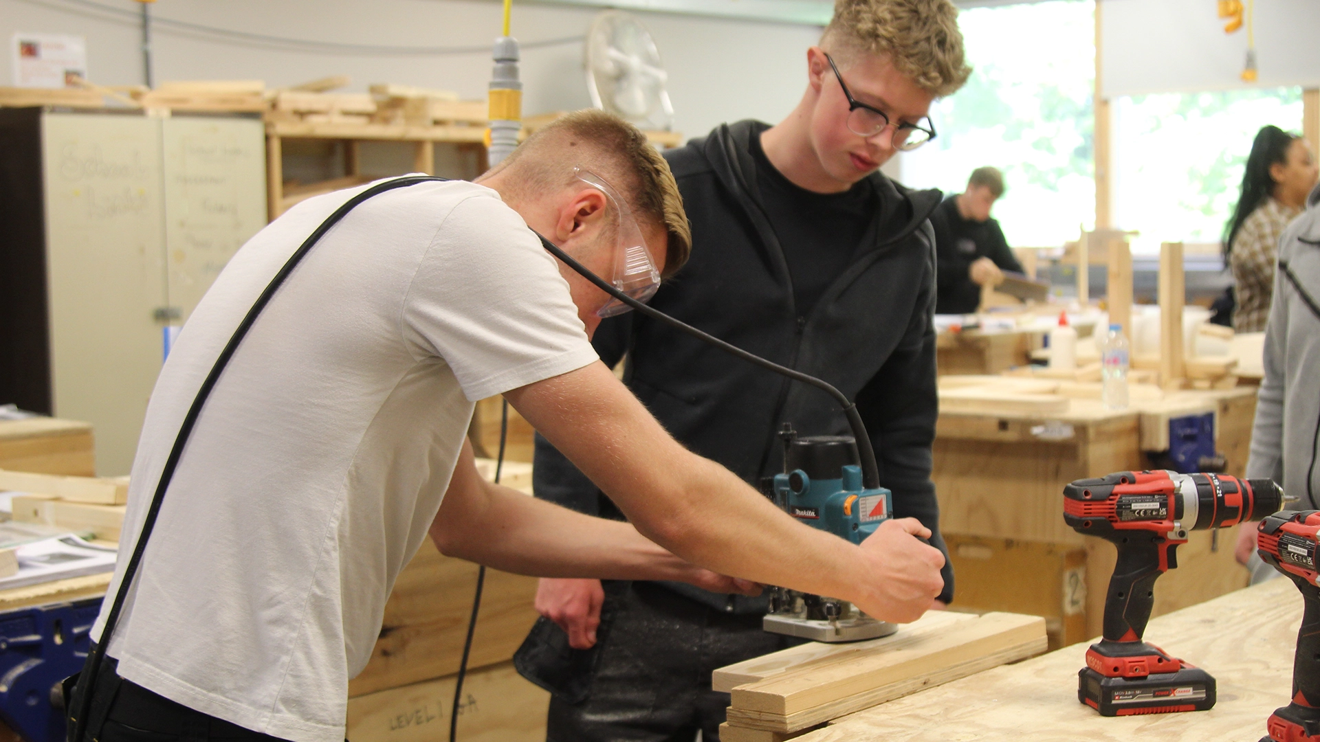 One person using a router on a wooden board while another observes in a carpentry workshop.