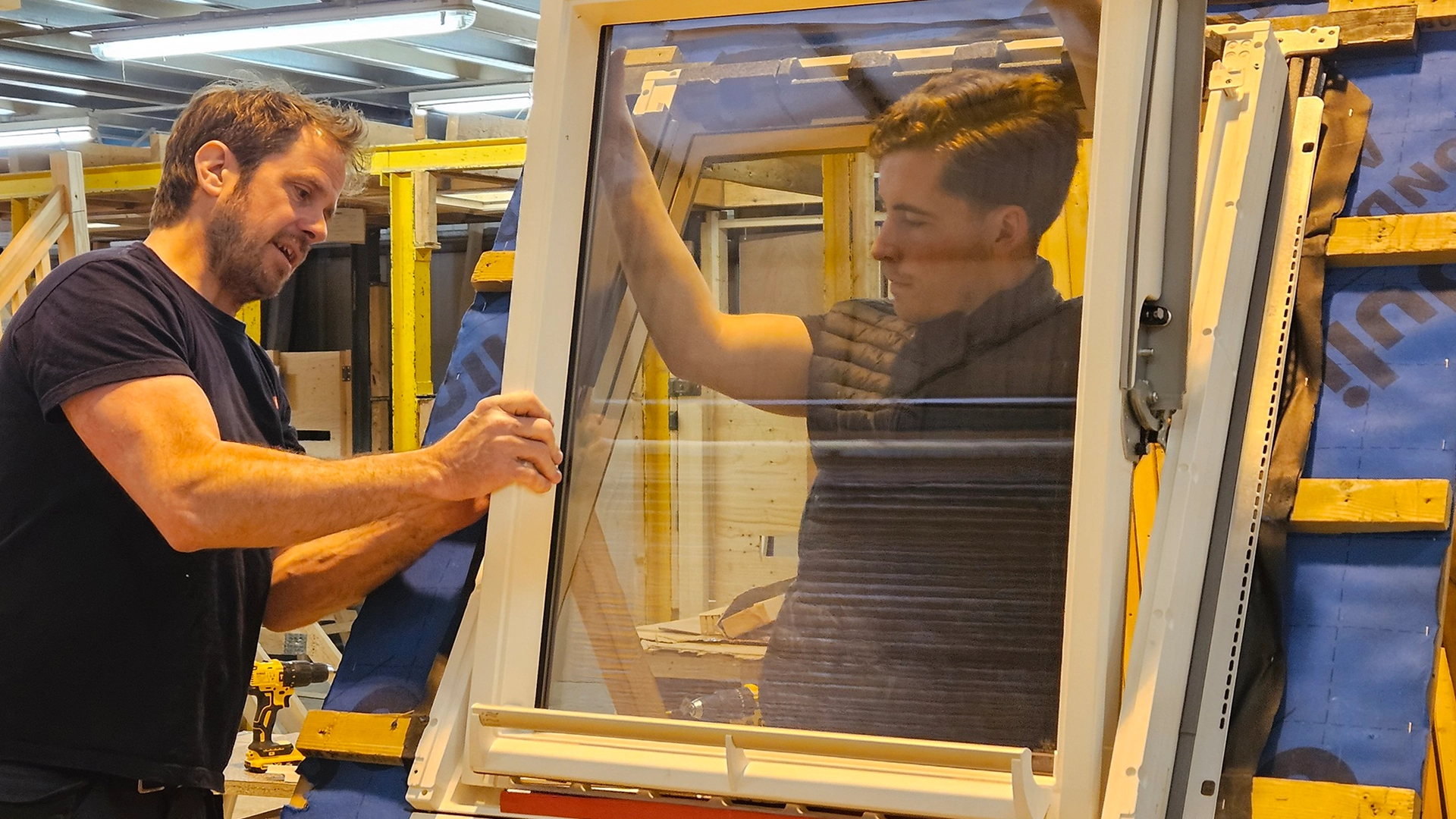 Two people installing or adjusting a window frame in a workshop environment.