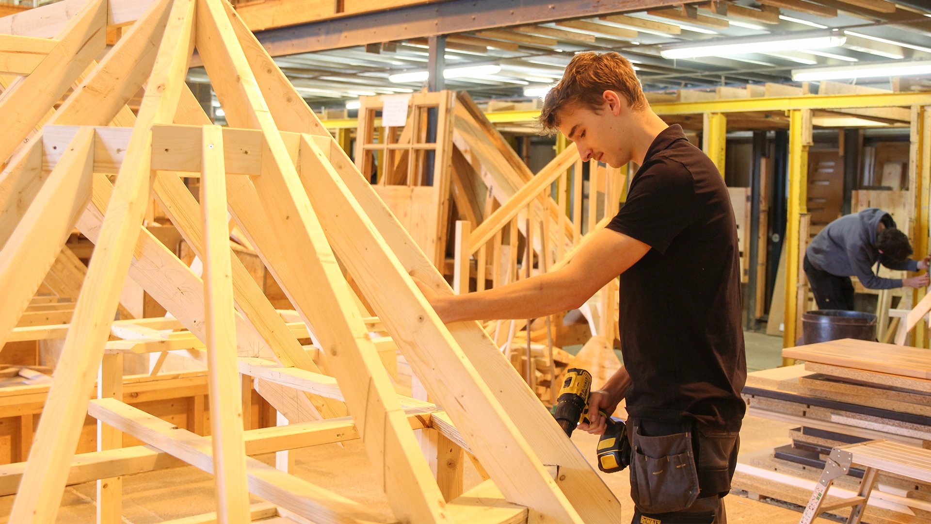 Person positioning timber components on a wooden frame structure while holding a drill in a workshop.