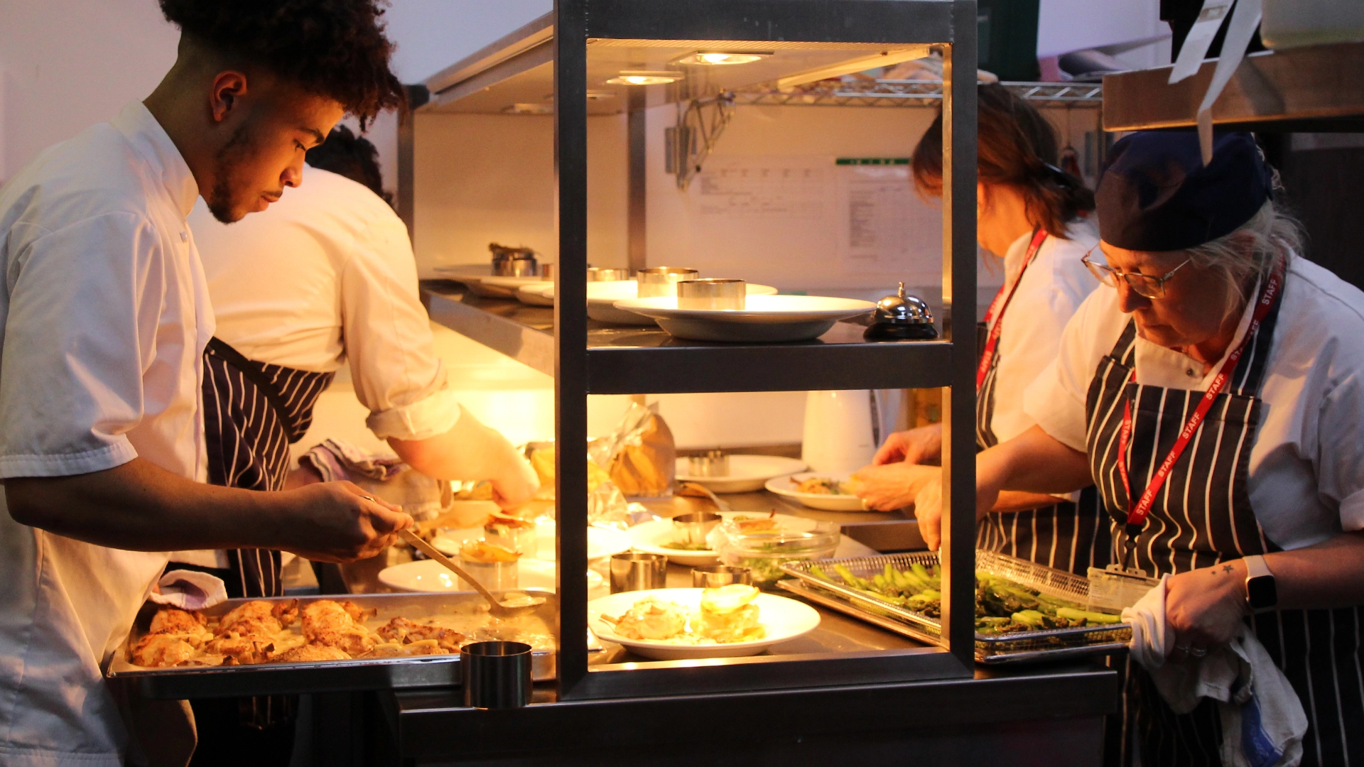 Students plating dishes under a heated pass in a professional training kitchen.