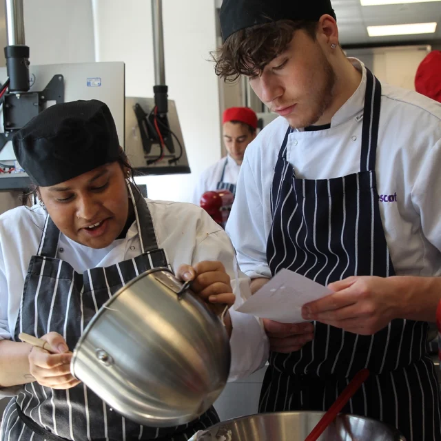 Students preparing ingredients together in a training kitchen, one pouring mixture while another checks a recipe.