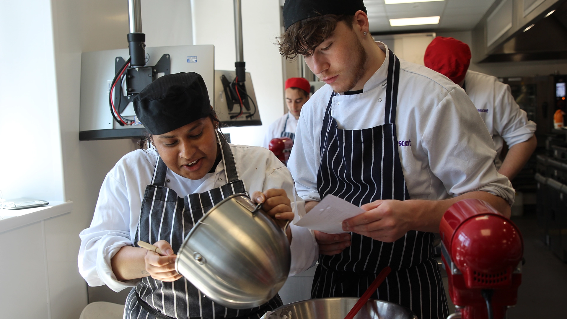 Students preparing ingredients together in a training kitchen, one pouring mixture while another checks a recipe.