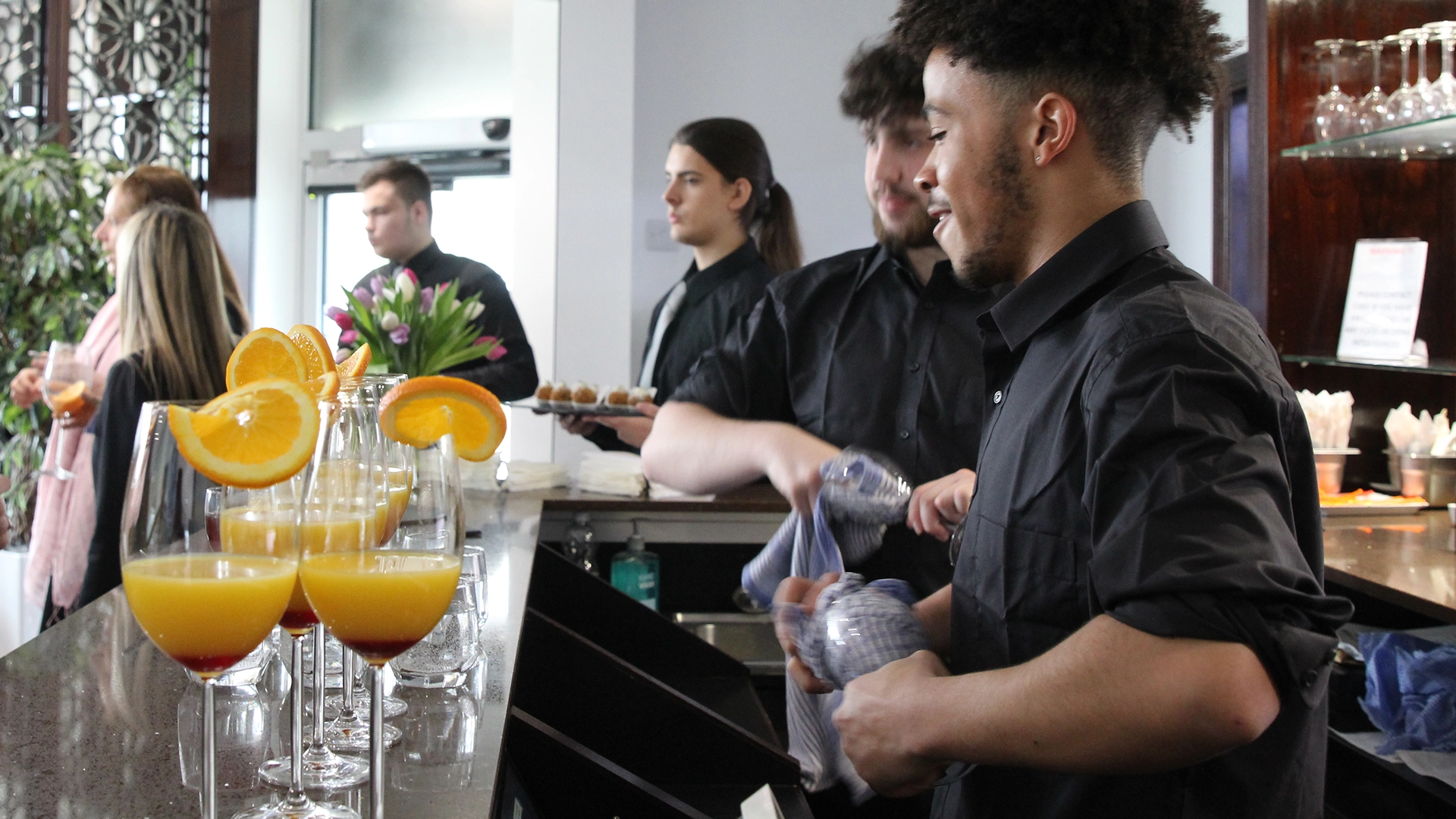 Student polishing glassware at a bar with orange drinks garnished with citrus slices.