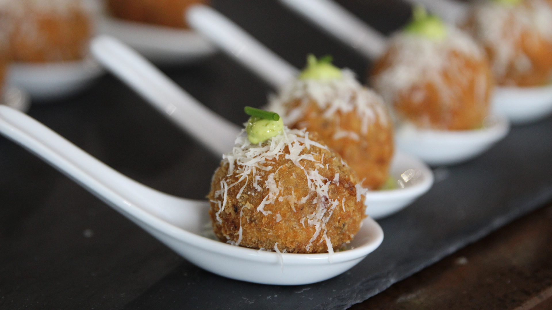 Close-up of fried arancini topped with grated cheese and green garnish on spoons.