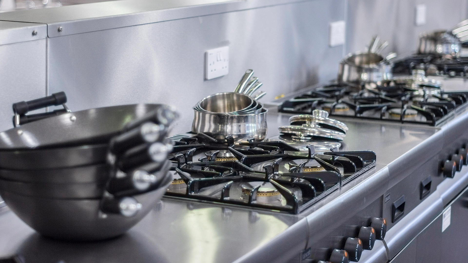 Row of stainless steel gas hobs with saucepans on a commercial kitchen worktop.