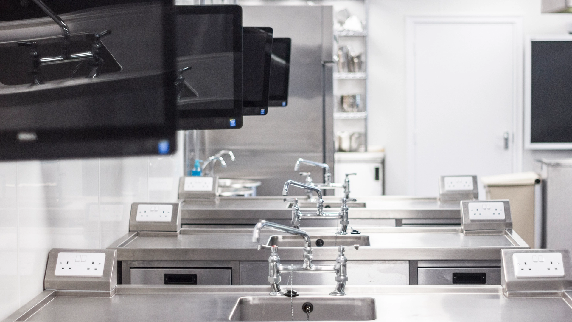 Stainless steel sinks with taps and mounted screens in a training kitchen.