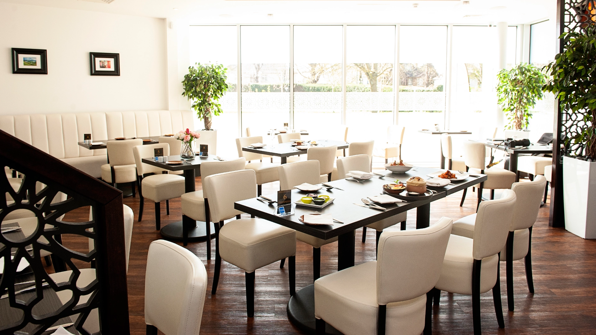 Bright restaurant dining area with neatly set tables, cream chairs and large windows letting in natural light.