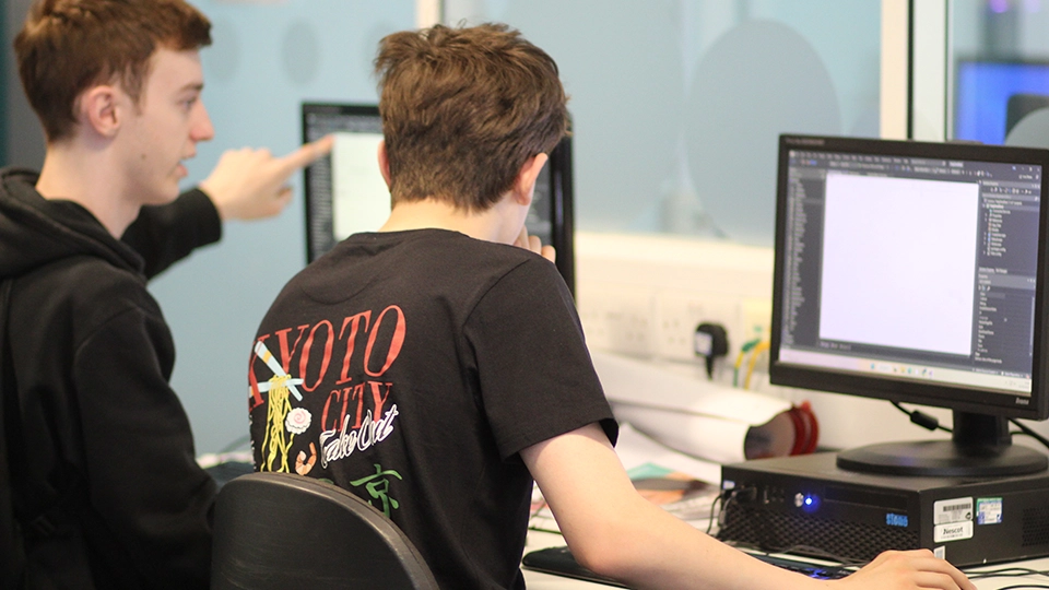 Student pointing at code on a monitor while another types at a computer
