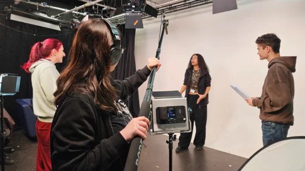 Students filming a scene in a studio, using a boom microphone, lighting equipment and script during a media production.