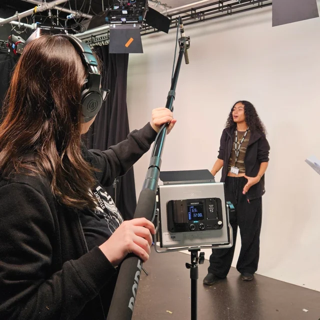 Students filming a scene in a studio, using a boom microphone, lighting equipment and script during a media production.