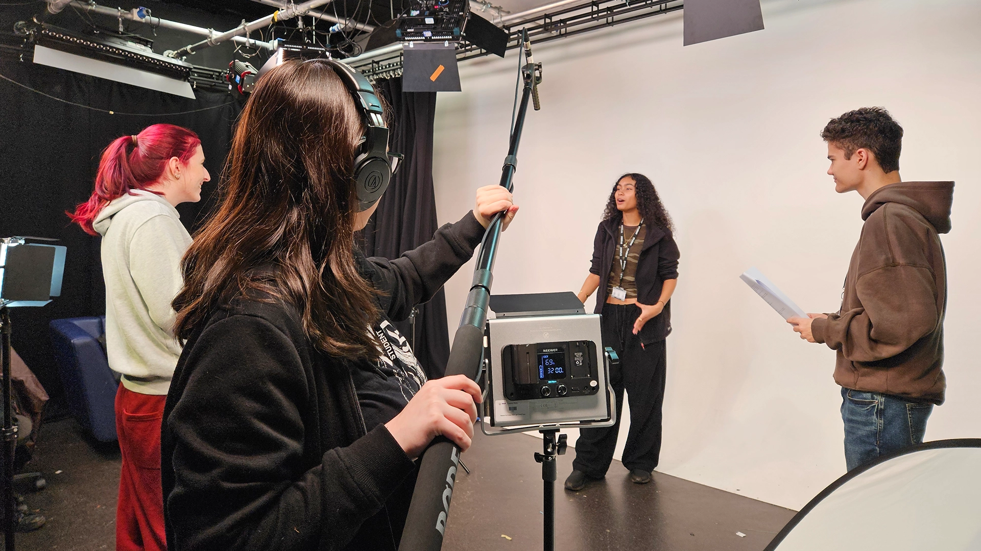 Students filming a scene in a studio, using a boom microphone, lighting equipment and script during a media production.