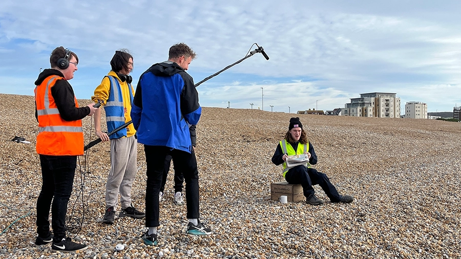 Students recording audio outdoors on a beach using a boom microphone.