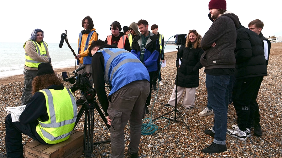 Group of students filming on a pebbled beach using cameras and sound equipment.