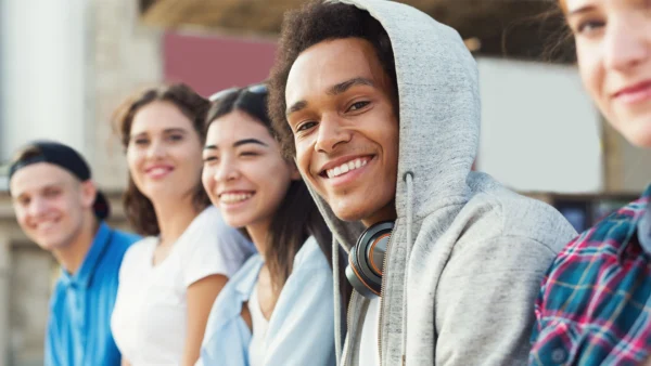 Group of students sitting outdoors in a row, one in a hoodie with headphones, others blurred in background.