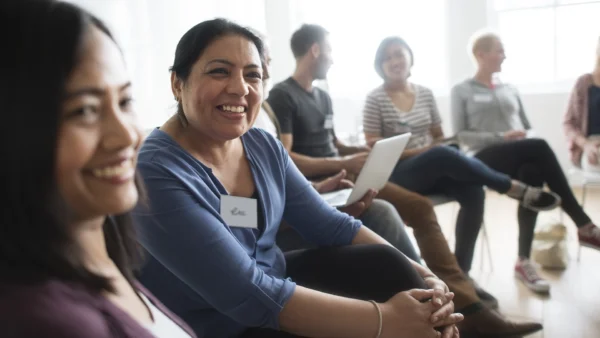 Adult learners seated in a circle in a classroom, one holding a tablet while others listen in a group discussion.
