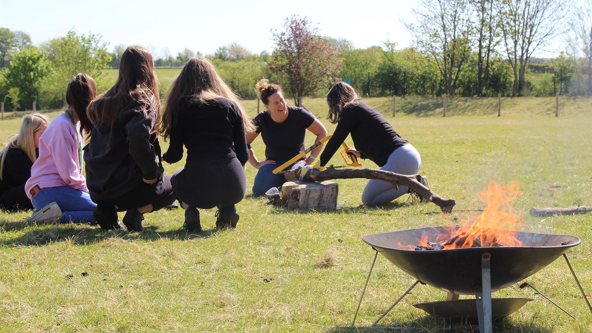 Group participating in outdoor activity, sawing wood near a small fire.