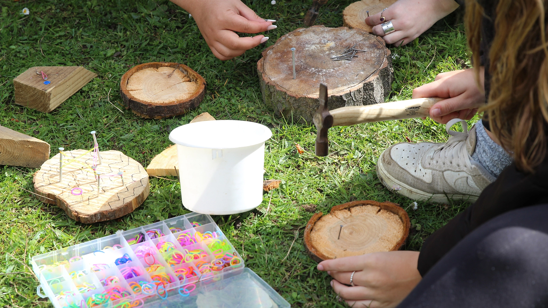 Hands using tools to hammer nails into wood outdoors on grass.