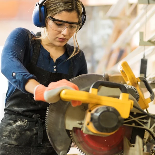 Person wearing safety goggles, ear defenders and gloves using a circular saw to cut wood in a workshop.