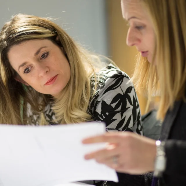 Adult learners reviewing printed materials together at a table, with others working in a classroom background.