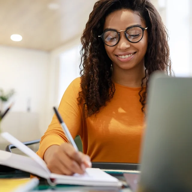 Student writing in a notebook while using a laptop at a desk in a bright study space