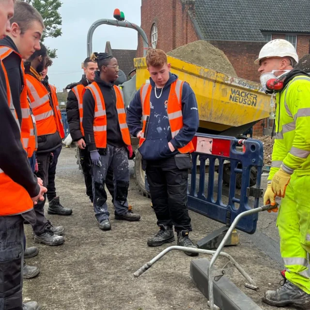 Students in high-visibility vests gather around an instructor demonstrating equipment at a roadside construction site near residential buildings.