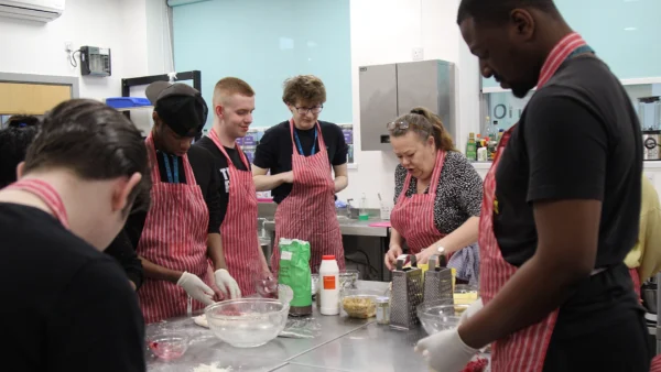 Students wearing aprons and gloves prepare food together in a kitchen classroom, with ingredients, bowls and utensils on a work surface.