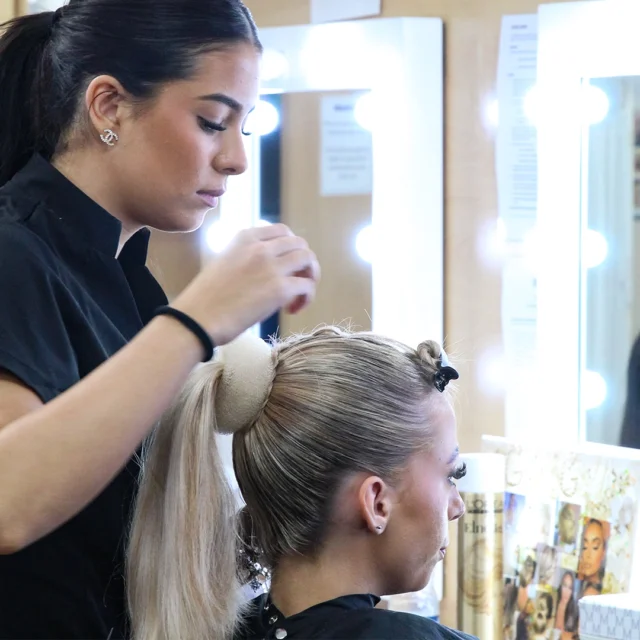Learner styles another learner’s hair into a high ponytail in a salon training room with illuminated mirrors and styling tools visible.
