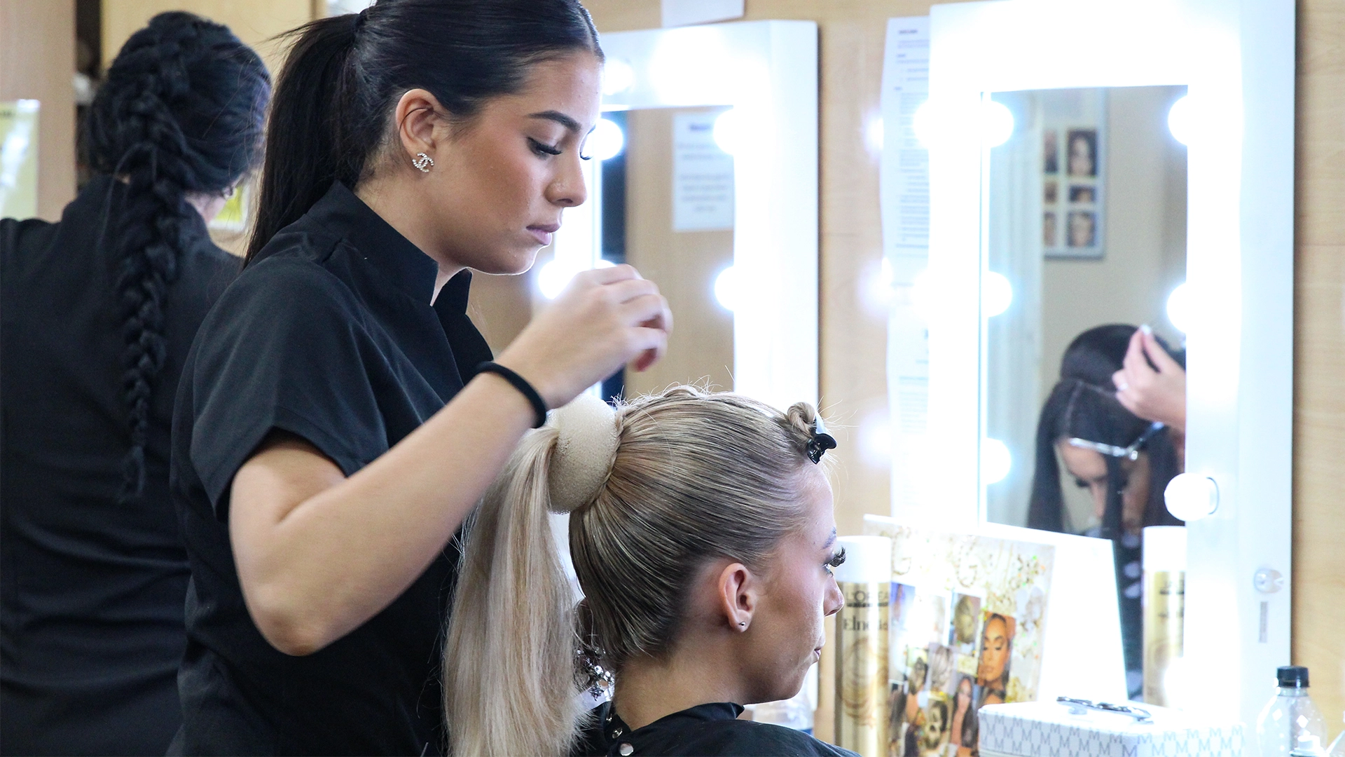 Learner styles another learner’s hair into a high ponytail in a salon training room with illuminated mirrors and styling tools visible.