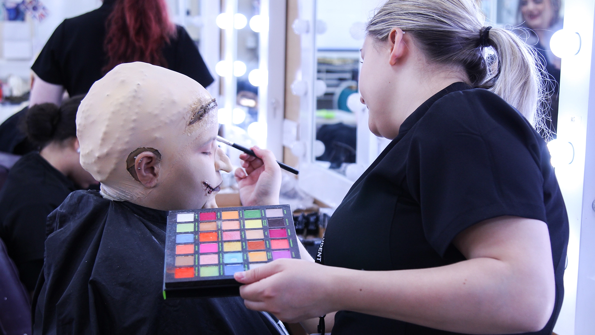 Student applying special effects make-up to a model wearing a prosthetic bald cap.